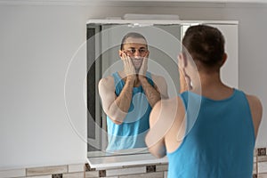 Male touching his beard and face. Sleepy man in bathroom looks at himself in mirror. Morning routine