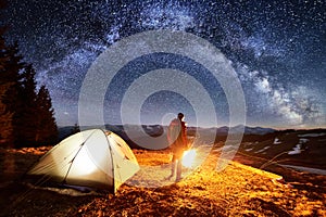 Male tourist have a rest in his camp near the forest at night. Man near campfire and tent under night sky full of stars