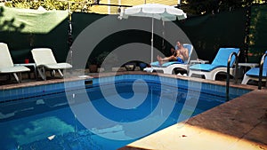 A male tourist is having rest on lounge chair near the swimming pool chatting to someone