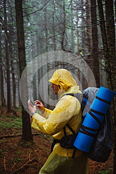 A male tourist in a raincoat and with a backpack on his back stands with a smartphone in his hand standing in the woods during a