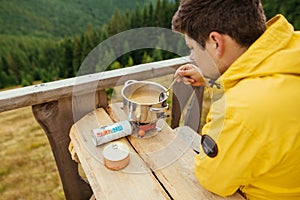 Male tourist sits at a table on the terrace of a country house in the mountains and prepares to eat on a burner on a background of