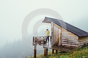 Male tourist standing on the terrace of a wooden house and drinking tea looking at the beautiful mountain views in foggy rainy