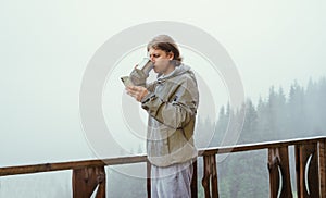 Male tourist stands in the morning on the terrace of a cottage in the mountains with a smartphone in his hands and drinks tea on a