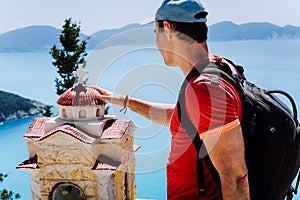 Male tourist touches thoughtful to small Hellenic shrine Proskinitari, Greece. Amazing sea view in the background