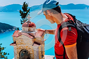 Male tourist touches thoughtful to small Hellenic shrine Proskinitari, Greece. Amazing sea view in the background