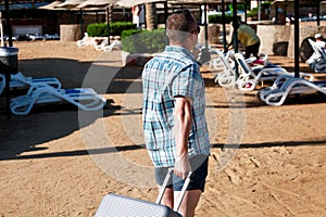 Male tourist and traveler walking and pull his traveled suitcase on sandy beach of hotel resort. Handsome man on vacation.