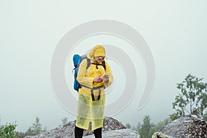 Male tourist in a yellow raincoat stands on top of a mountain in rainy weather against a background of clouds and uses a