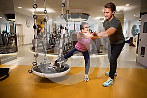 Male trainer helping mature caucasian woman doing sport exercise on hanging mat