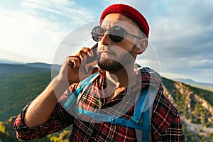 A male traveler in a red hat with a backpack is talking on the phone against the background of mountains.