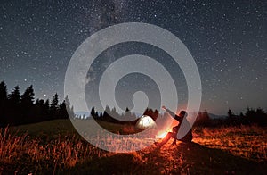 Male traveler sitting near campfire and pointing at stars.