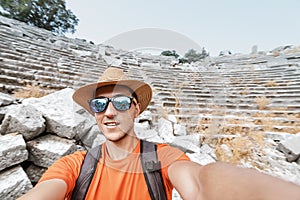 Male traveler wearing sunglasses and a hat takes a selfie against the ruins of an ancient amphitheater in the Termessos National