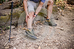 A male trekker sits on a wooden log and massages his leg, suffering from knee pain, leg cramping
