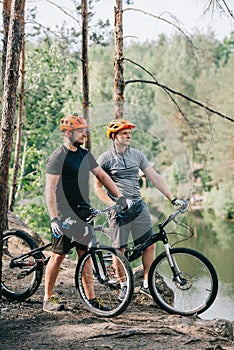 male trial bikers in helmets with mountain cycles resting with sport bottles of water near river