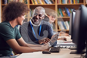 Male University Or College Student Working At Computer In Library Being Helped By Tutor