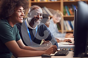 Male University Or College Student Working At Computer In Library Being Helped By Tutor