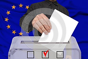 Male voter drops a ballot in a transparent ballot box against the background of the European Union national flag, concept of state