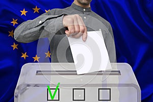 Male voter drops a ballot in a transparent ballot box against the background of the European Union national flag, concept of state