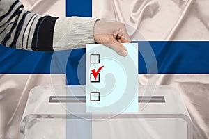 Male voter drops a ballot in a transparent ballot box against the background of the national flag of Finland, concept of state