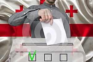 Male voter drops a ballot in a transparent ballot box against the background of the national flag of Georgia, concept of state