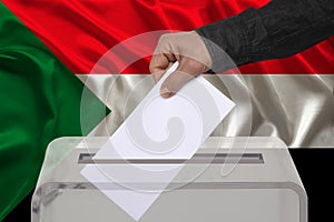 Male voter drops a ballot in a transparent ballot box on the background of the national flag of Sudan, concept of state elections