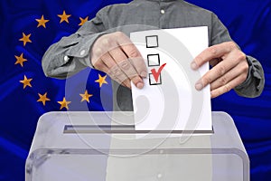 Male voter drops a ballot in a transparent ballot box against the background of the European Union national flag, concept of state