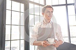 A male waiter in a gray apron holds out a cup of coffee. The barista serves a cup of hot coffee, against the background