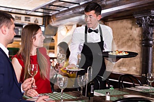 Male waiter is serves an order for smiling couple who is drinking wine and eating