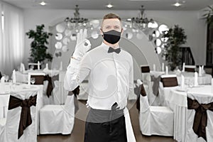 A male waiter in a white shirt and a protective medical mask in the restaurant hall shows the ok sign