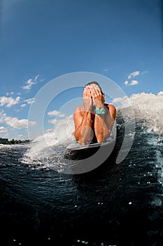 Male wakesurfer rides wave while lying on surfboard covering his face with hands.