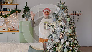 Male waving hand near Xmas tree. Happy young man in warm clothes and Santa hat smiling and waving hand while standing