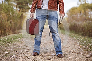 Male wearing a leather jacket and jeans standing on a pathway holding old suitcase and bible