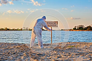 Male wearing protective suit stands with his back near warning sign with inscription beach closed along the sand beach