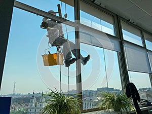 A male window washer worker, industrial climber hangs on a tall building, skyscraper and washes large glass windows for