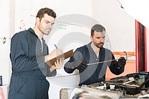 Male Worker Checking Motor Oil While Coworker Writing In Clipboard