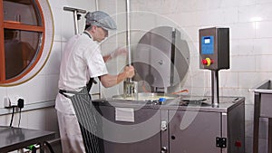 Male worker in gloves mixing and cutting pieces of a soft cheese in the vat with a big instrument at the factory