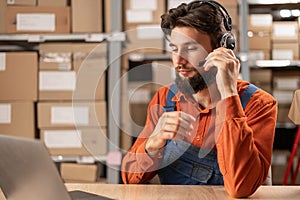 Male worker with headset working in on-site office of a warehouse using laptop computer consulting clients