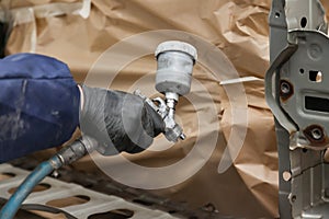 A male worker in jumpsuit and gloves paints with a spray gun a front frame part of the car body in black after being damaged at an