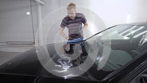 Male worker polishing black car hood with a dry towel after waxing it on a carwash.