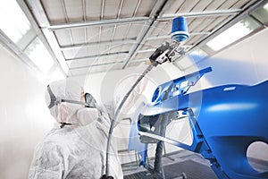 Male worker in protective clothes and mask painting car bumper using spray paint.