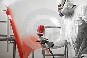 Male worker in protective clothes and mask painting hood of car using red spray paint.