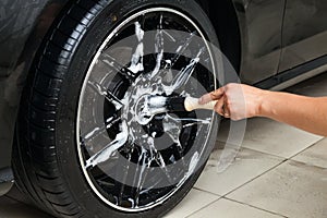 A male worker washes a black car with a special brush for cast wheels and scrubs the surface to shine in a vehicle detailing