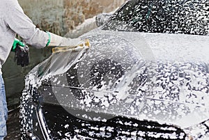 Male worker washing car at car station outdoors with foam and yellow sponge.