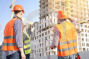 Male workers engineers at construction site in helmets hardhats look at high-rise building skyscraper