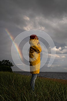 Male in yellow raincoat wear red hat standing on the beach in rainy weather, looks at dramatic cloudy sky and sea, rainbow on