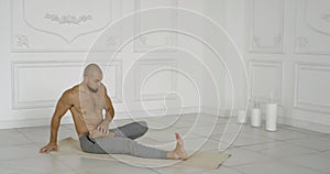 Male yoga follower is performing stretching of legs sitting on floor in big light hall