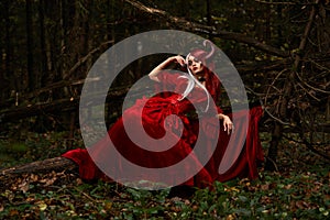 Maleficent Woman in Red Clothing and Horns in dark Forest. Posing in magik forest