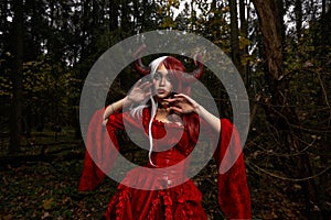 Maleficent Woman in Red Clothing and Horns in dark Forest. Posing in magik forest