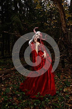 Maleficent Woman in Red Clothing and Horns in dark Forest. Posing in magik forest