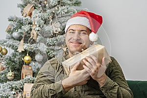 A man of 30-35 years of oriental appearance in a red hat is holding a gift box against the background of a Christmas tree.