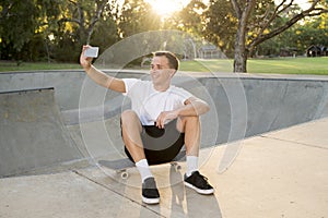 Man 30s sitting on skate board after sport boarding training session taking selfie photograph portrait or picture on mobile phone